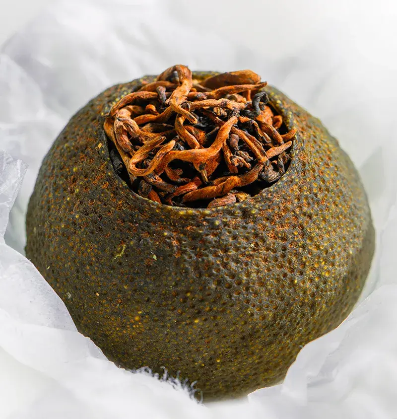 Close-up view of a whole dried Xiao Qing Gan citrus fruit stuffed with aged Pu-erh tea leaves. Its textured peel and golden tea buds are clearly visible.